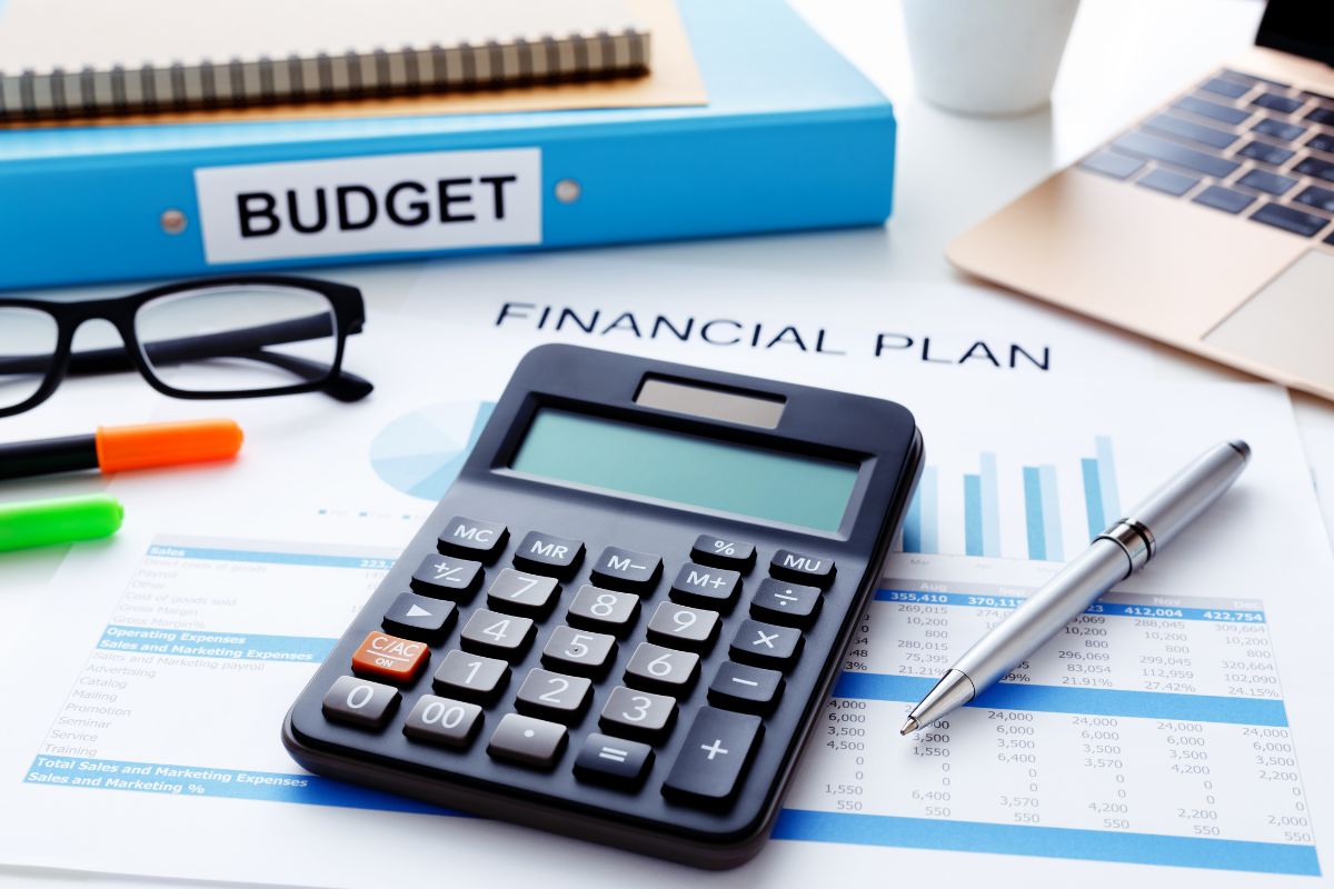 The image tells about a laptop, notebooks, files, an eyeglass, pens, a calculator, and a piece of paper with notes and graphs referring to regularly reviewing and adjusting your financial plan keeping it updated, and helping you stay in control.