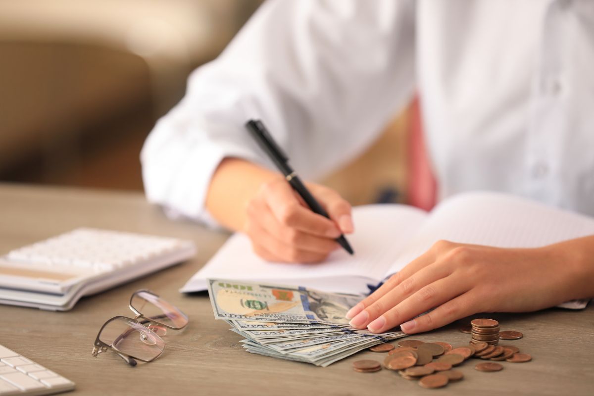 The image shows an eyeglass, a calculator, coins, and a girl's hand and writing on the notebook while holding dollar bills which means that setting clear financial goals helps you make smart money choices and avoid wasteful spending.