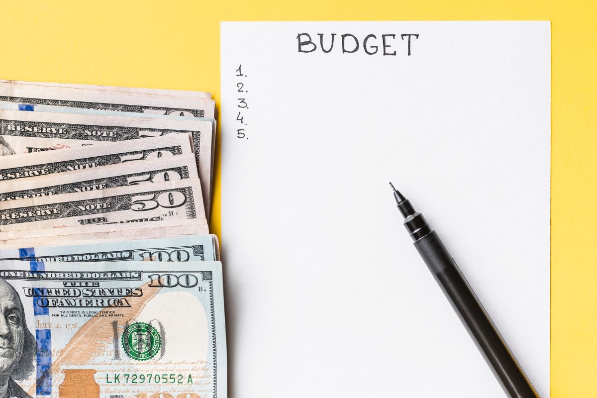 The photo shows dollar bills, a pen, and a piece of paper with notes indicating that creating and sticking to a budget is a simple way to control your money instead of letting it control you. 