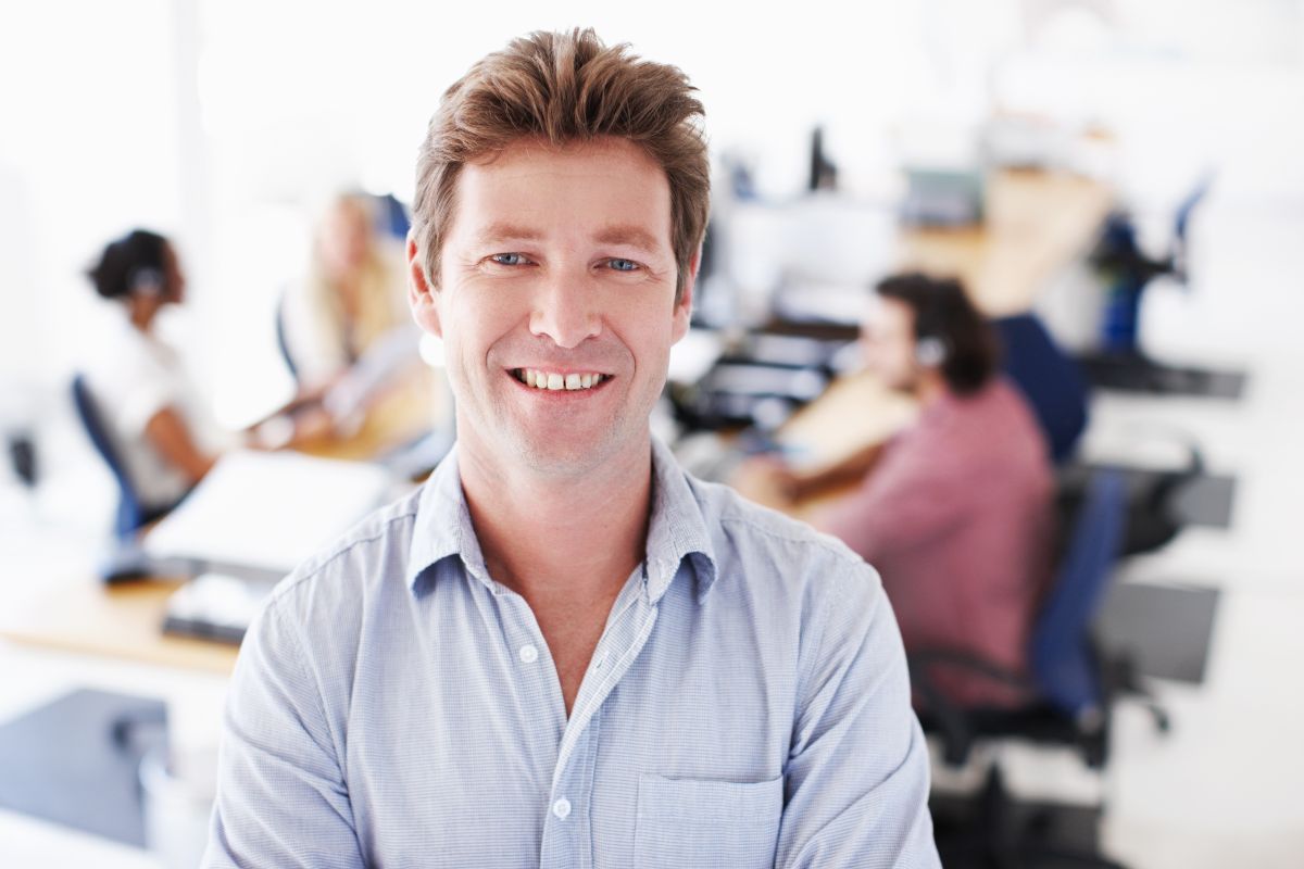 The photo shows a group of people working at the back and a man standing in front and smiling which means developing a strong work ethic and discipline helps keep you more that leads to financial security later.