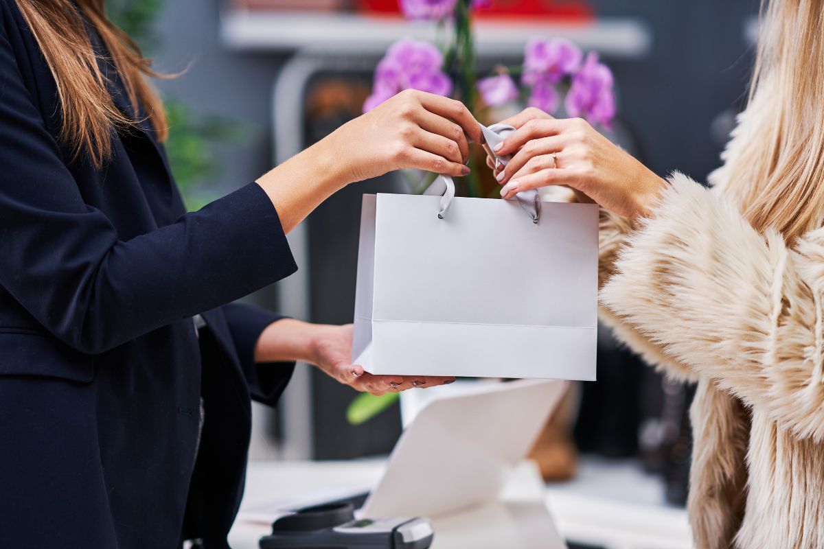 The image shows a swiping machine and a cashier handing a shopping bag to the customer's hand referring to avoiding lifestyle inflation leading to wealth in the future.