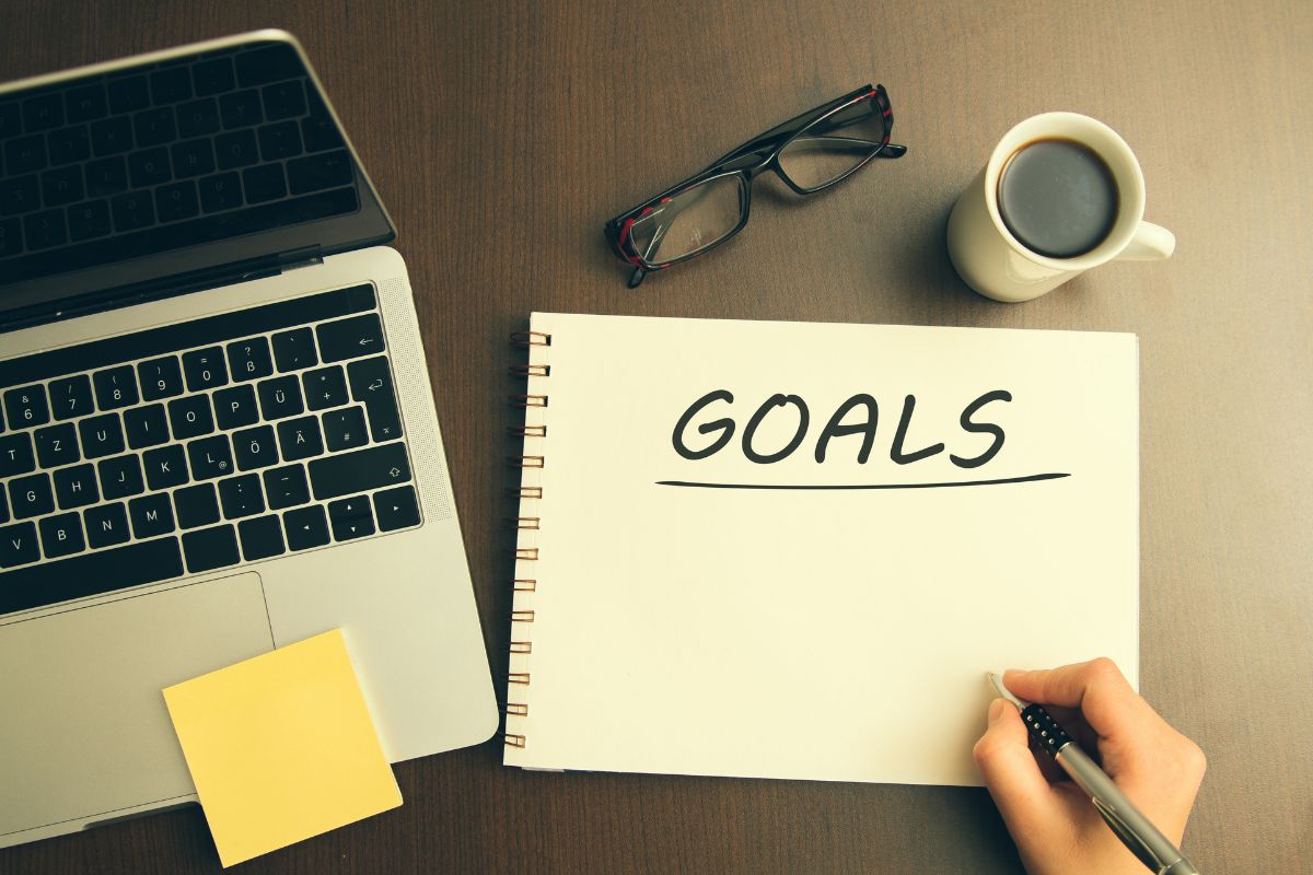 The photo shows a laptop, a coffee, an eyeglass, a sticker note, and a girl's hand, and writing on the notebook referring to setting your goal helps to remind yourself of your goal when the challenge gets tough.