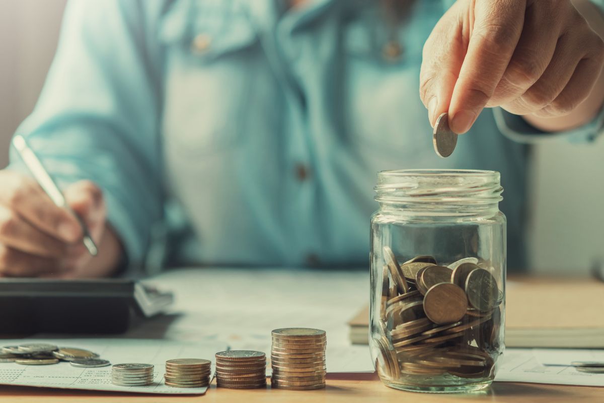 The photo shows stock coins, papers on the table, a calculator, and a girl's hand holding a pen while dropping a coin in a jar filled with coins referring to automate savings helps build the habit of putting money side and boosts financial growth.