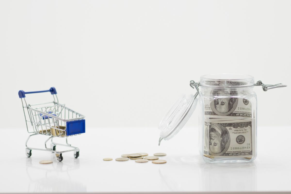 The picture shows coins, a mini cart, and a jar filled with dollar bills which means setting a strict budget before shopping makes it easier to stretch money and get the best value.
