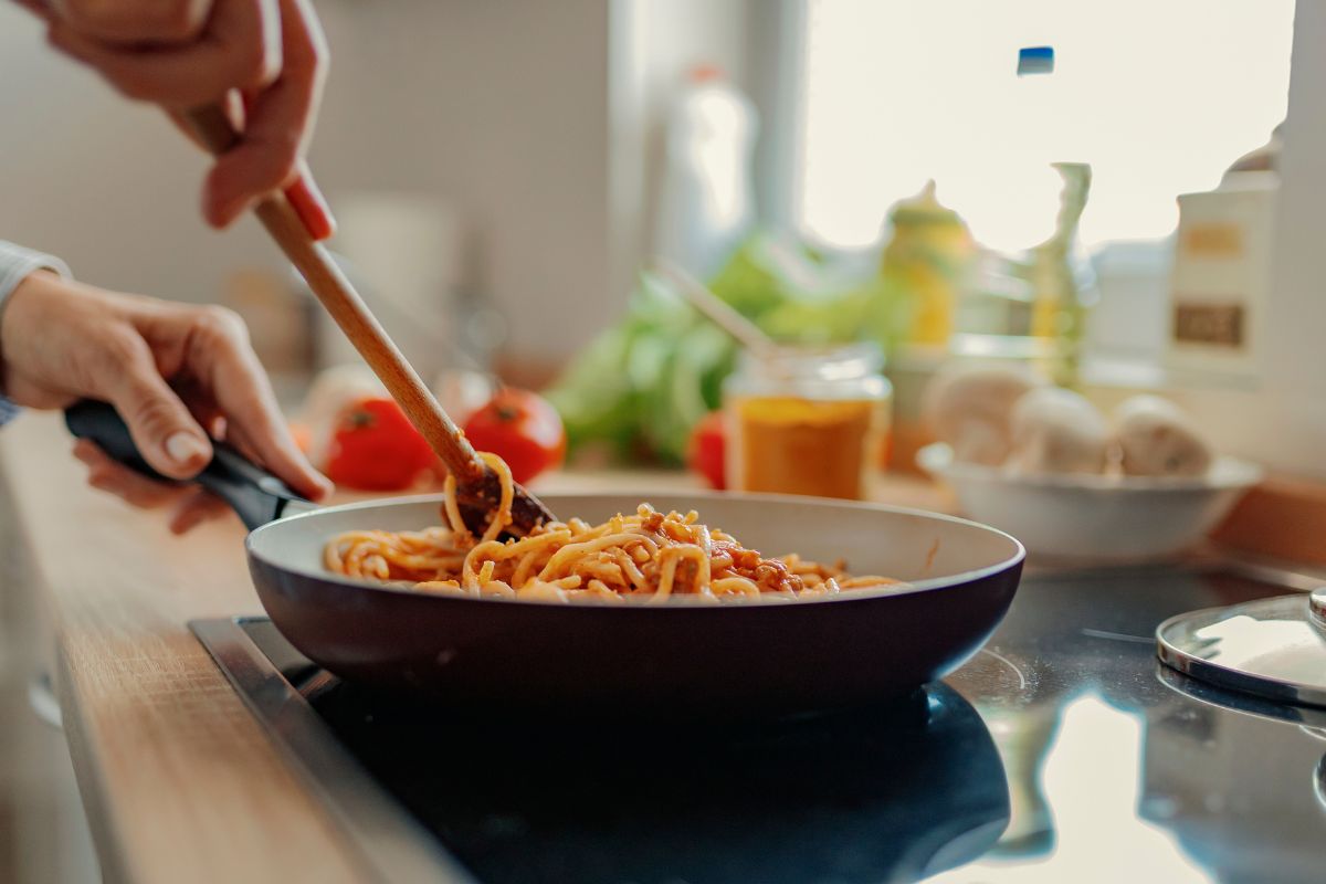 The image tells about ingredients, tomatoes, and a girl's hand stirring the pasta in a pan indicating that cooking at home instead of buying takeout meals makes staying on a budget easier.