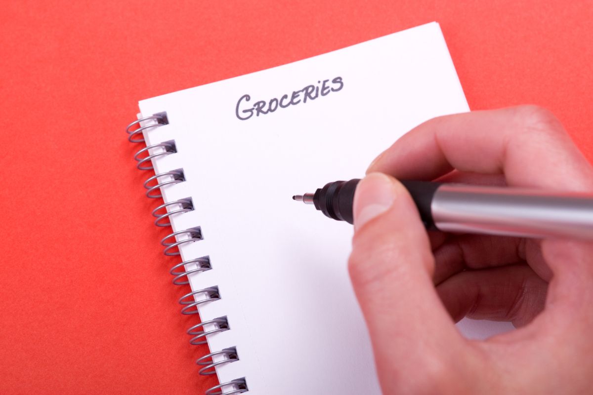 The photo shows a man's hand and writing on the notebook which refers to getting your copy of the budget grocery list to avoid impulse purchases and make thoughtful choices at the store.