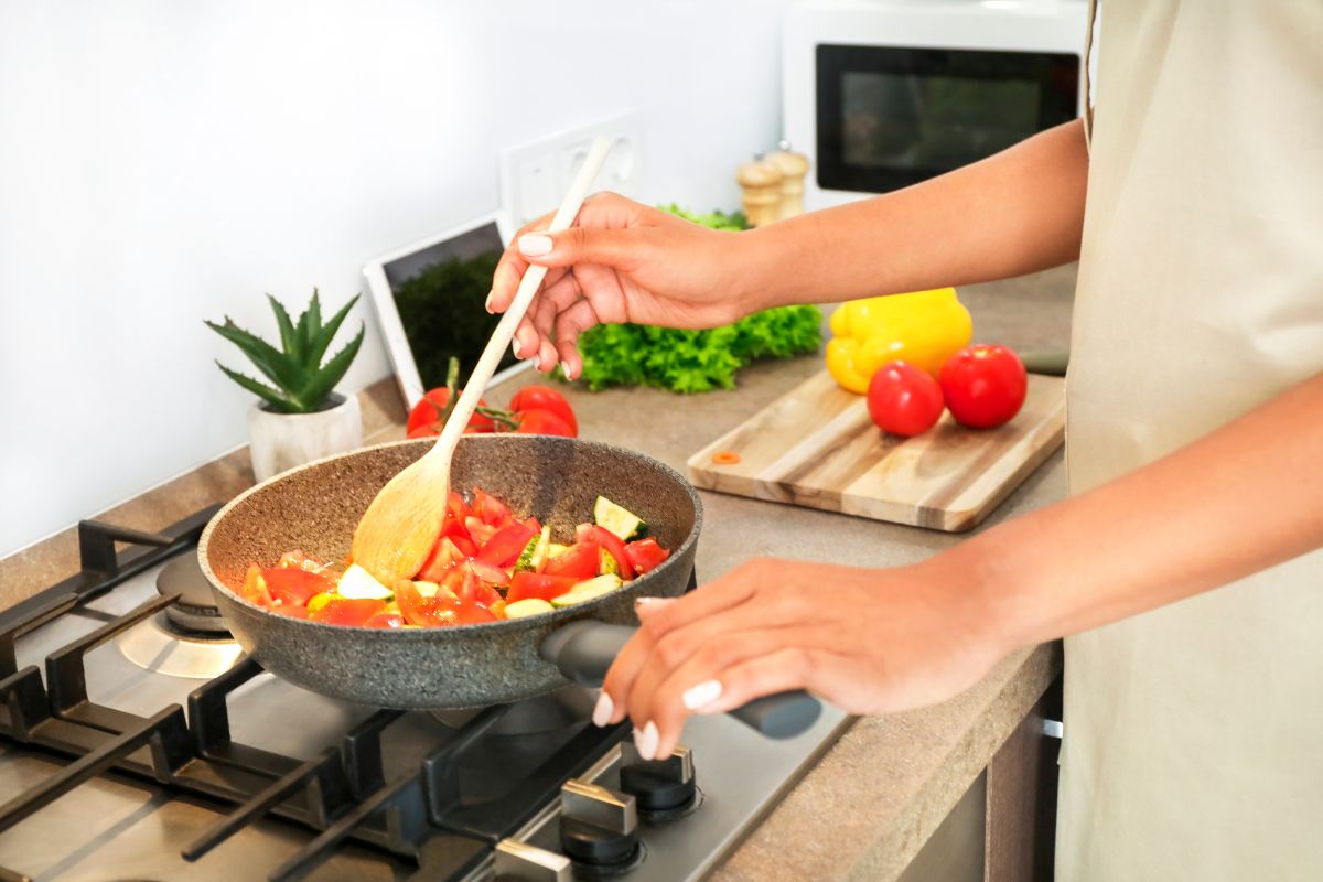 The photo shows ingredients, a plant, a tablet device, and a girl's hand stirring the vegetables in a pan which means cooking at home instead of eating out can stretch your budget and improve your diet.