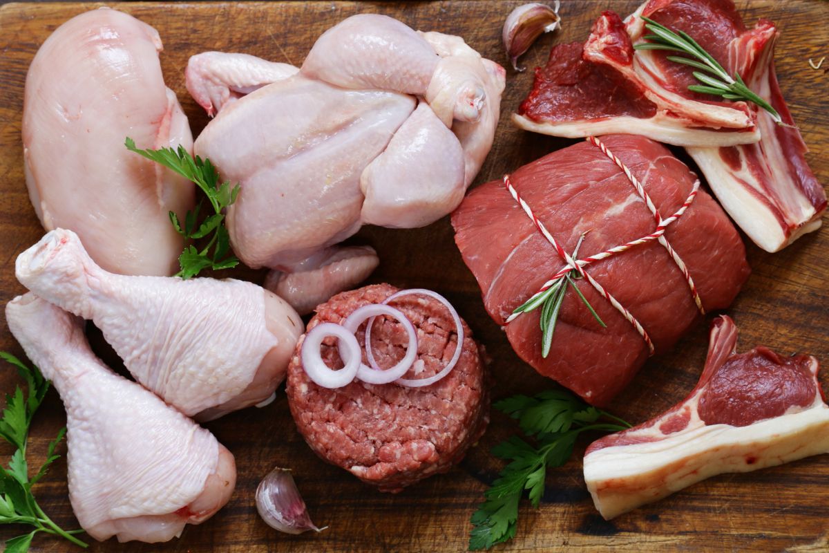 The photo tells about different kinds of meats on the wooden tables with leaves, onions, and garlic indicating that reducing meat consumption helps both your wallet and your health.