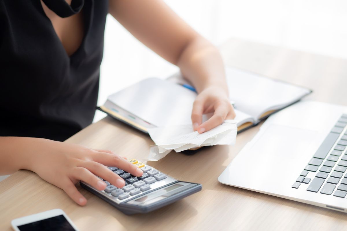 The image shows a laptop, a notebook, a phone, and a girl's hand holding receipts while calculating which means tracking expenses regularly helps you spot spending habits that might hold you back.  