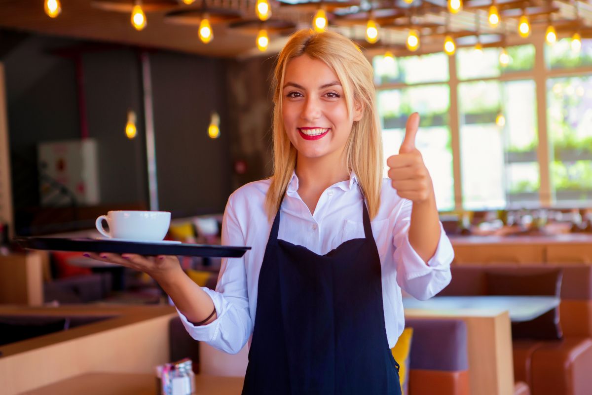 The image shows tables, chairs, and a girl holding a tray with a mug while doing an ok sign which means your child can still succeed through work and making the most of the opportunities available.