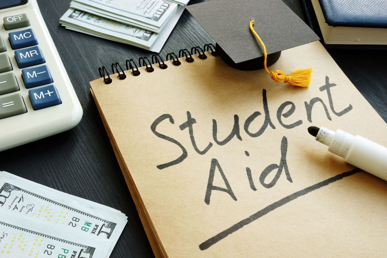 The picture shows dollar bills, a book, a marker, a mini graduation cap, and a notebook with notes indicating that many students pay their way and still thrive which can teach them valuable life skills.