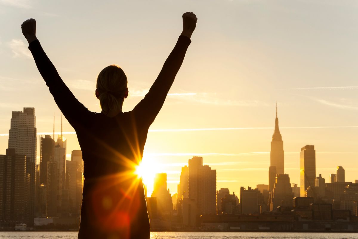 The picture of a girl raising both hands on the beautiful sunset view with building around which refers to many successful individuals who built their wealth through small and smart decisions made over time.