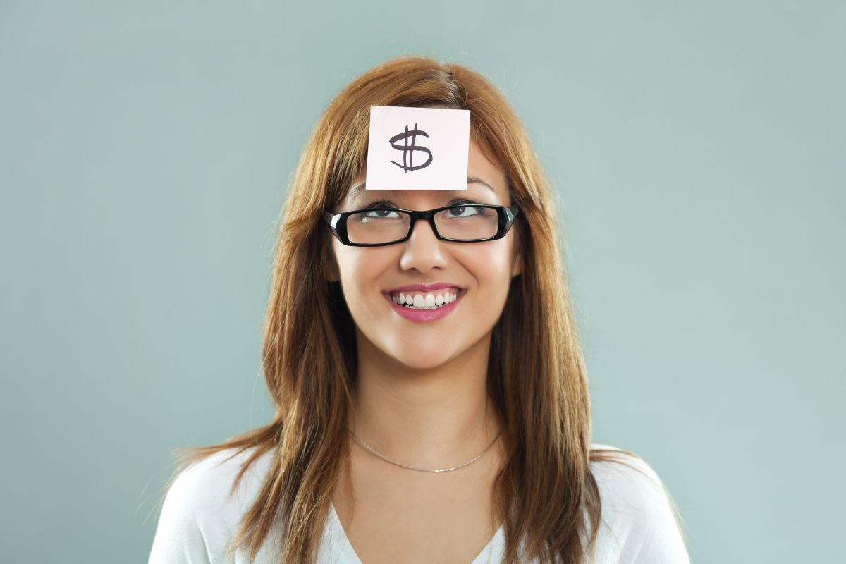The photo tells about a girl with a piece of paper on her forehead with a dollar sign indicating to time to manifest your money through positive financial habits starting with shifting your mindset and taking small, consistent actions.