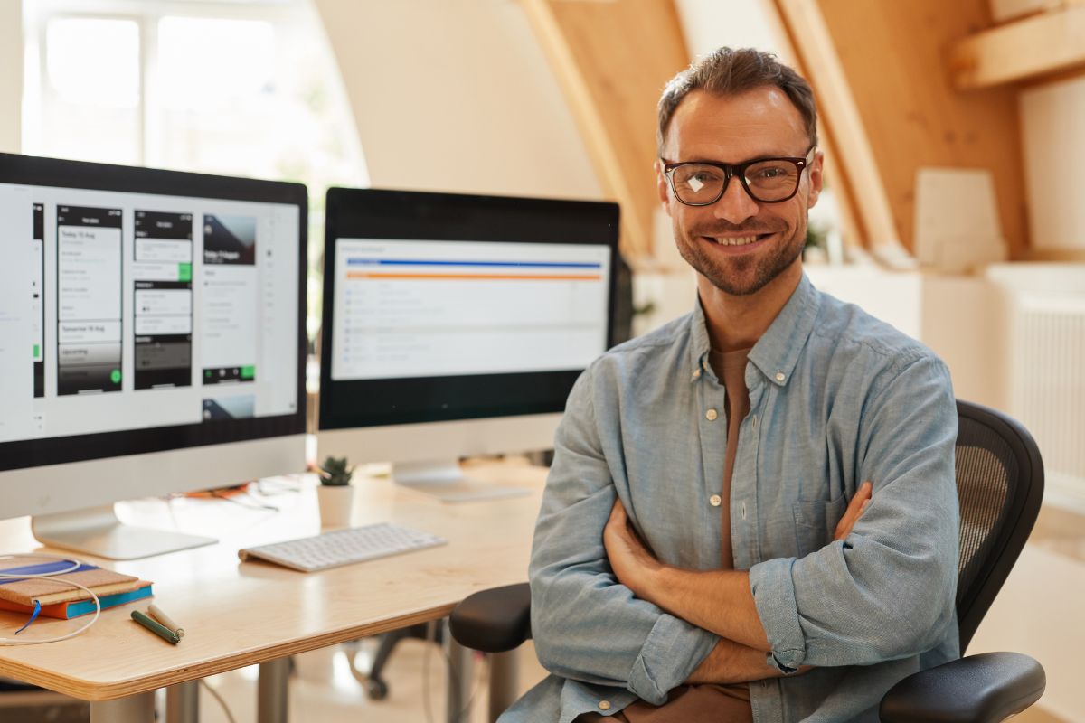 The image shows monitors with screen displays and a man sitting on a chair and smiling which refers to software engineer as one of the remote work that pay well.