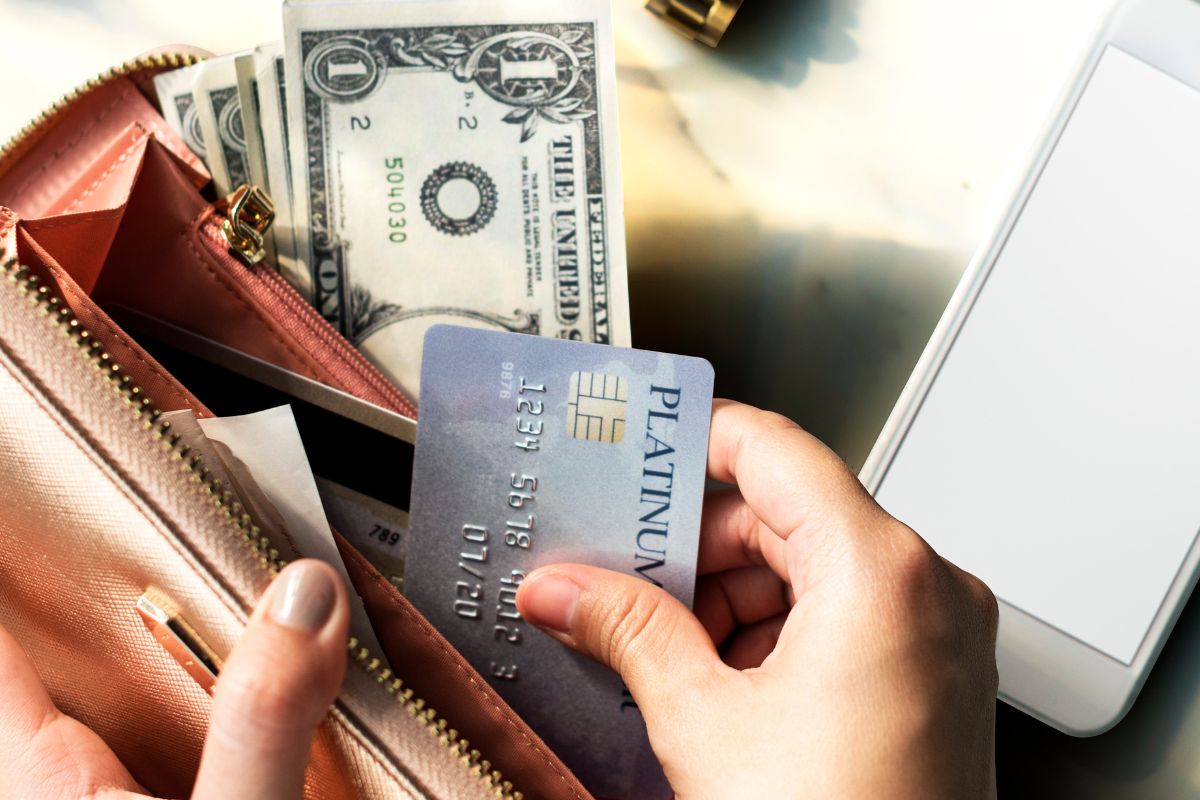The picture shows a phone and a girl's hand holding a wallet with dollar bills and putting back the card referring to how living below your means helps build long-term wealth.
