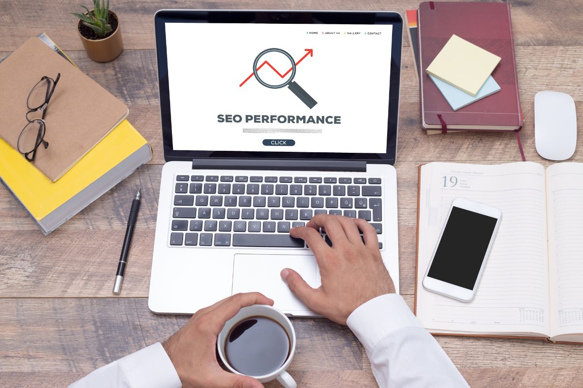 The photo shows books, an eyeglass, notebooks, a phone, a mouse, and a man's hand using a laptop with a screen display while having a coffee indicating that SEO Specialists help websites show up higher in search engine results and making it great for remote work.