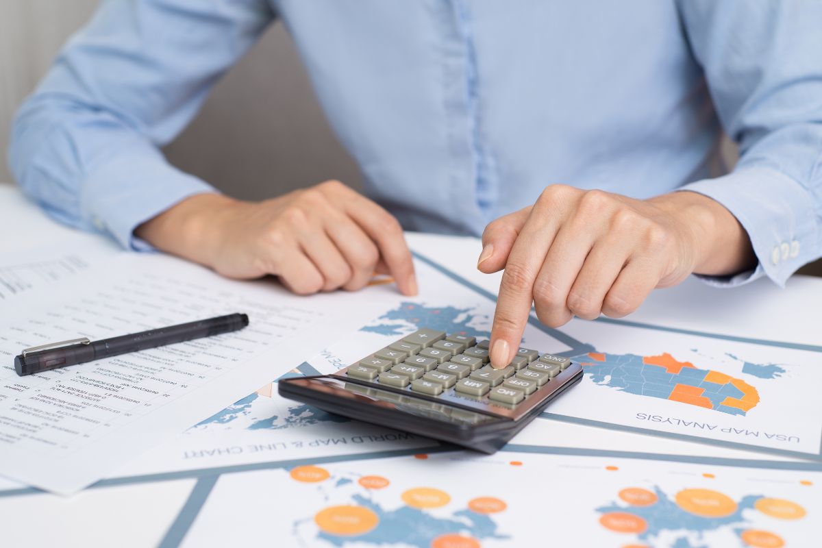 The photo shows papers with graphs, a pen, and a man's hand using a calculator which refers to financial analysts helping businesses understand their money and a strong remote career.