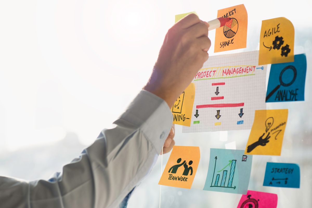 The image tells about paper with icons, notes, and a man's hand holding a marker referring to a project manager who keeps teams on track to complete tasks and is a good online job source.