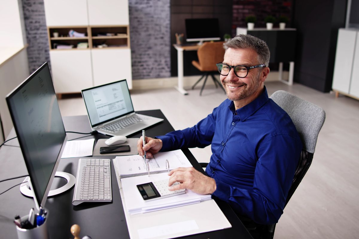The picture tells about a computer set, a laptop, a mouse, a phone, and a man sitting while writing on the papers and using a calculator which refers to a virtual bookkeeper managing financial records for businesses and could be a solid career choice.