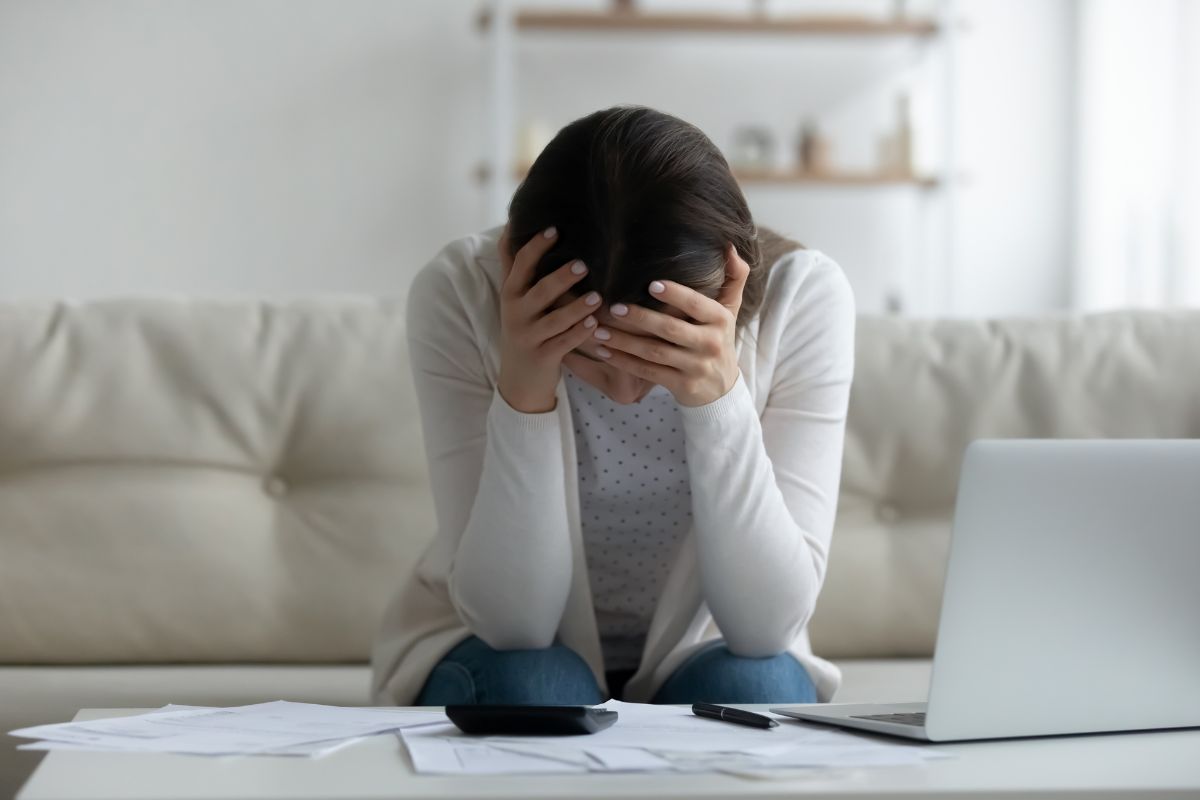 The photo of papers on the table, a calculator, a pen, a laptop, and a frustrated girl sitting on the couch which means no spend a month reduces financial stress that can be a big step toward financial security.
