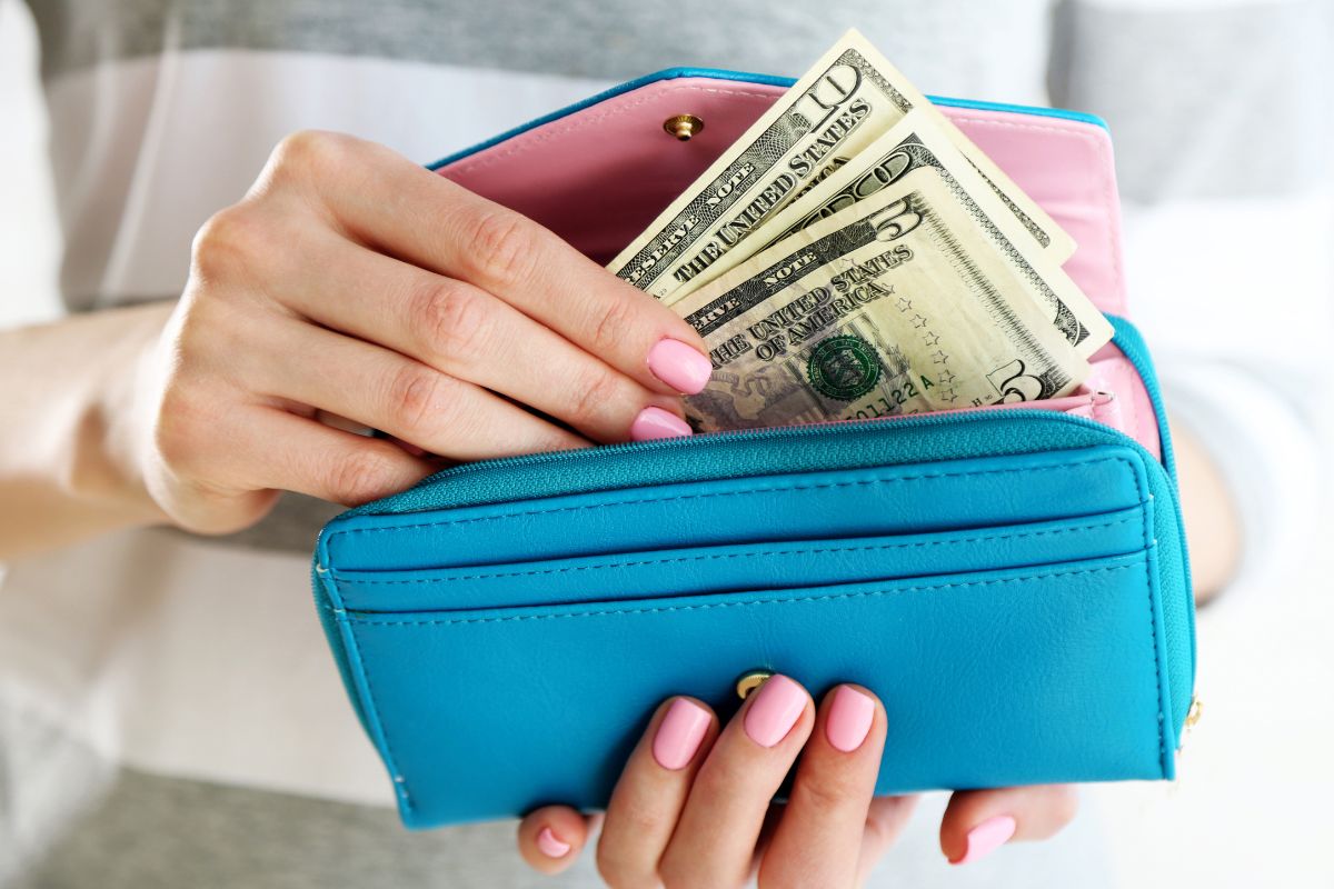 The photo of a girl's hand putting the dollar bills inside her wallet refers to starting your no-spend challenge and making it easier to stick to a budget and reach financial goals.