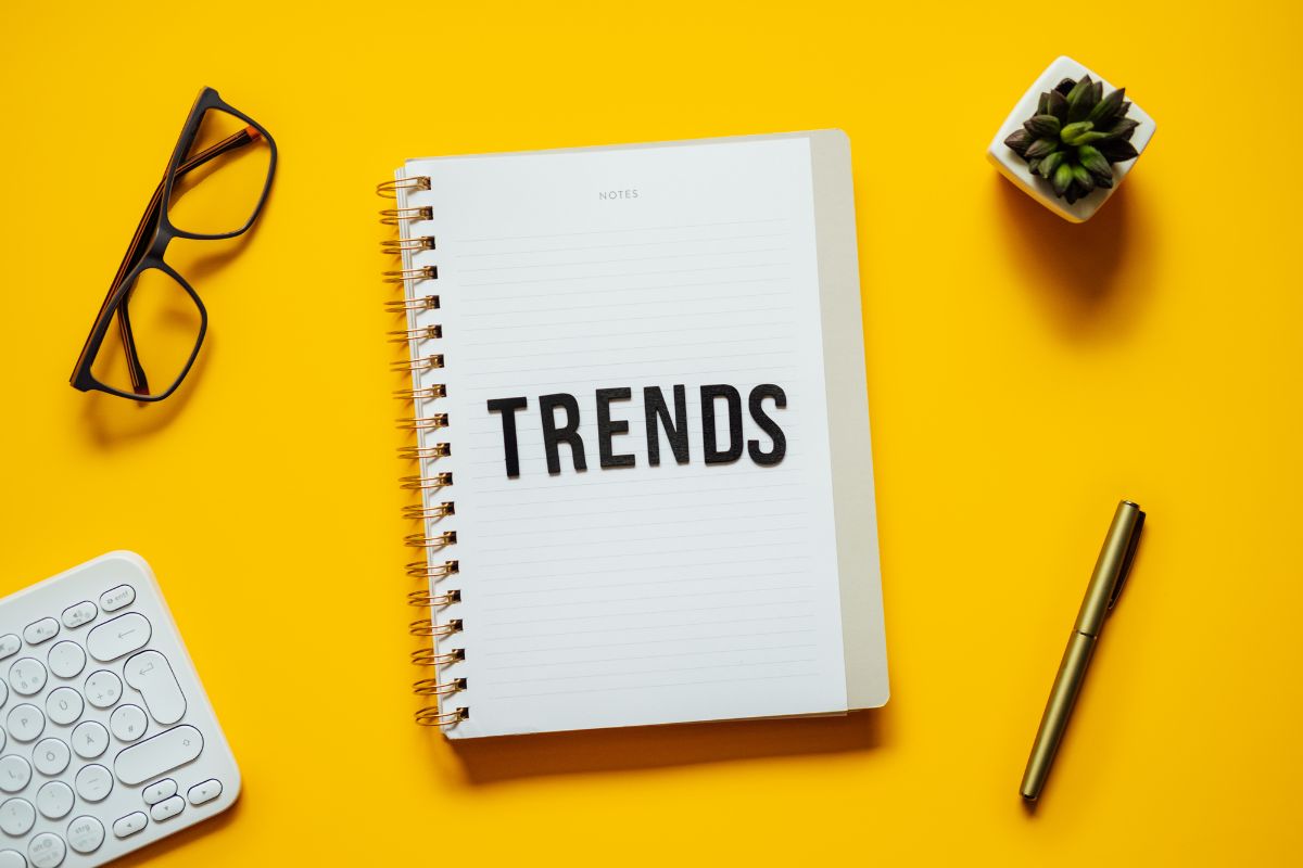 The photo shows an eyeglass, a plant, a keyboard, a pen, and a notebook with a note referring to research trending making it easier to turn a profit.