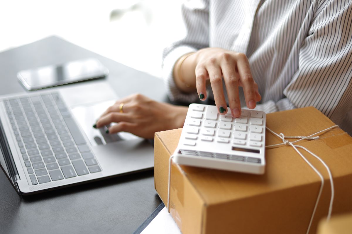 The image tells about a box, a phone, and a girl's hand using a laptop while calculating referring to the factor in shipping costs keeps pricing accurate and ensures every sale is worth it.
