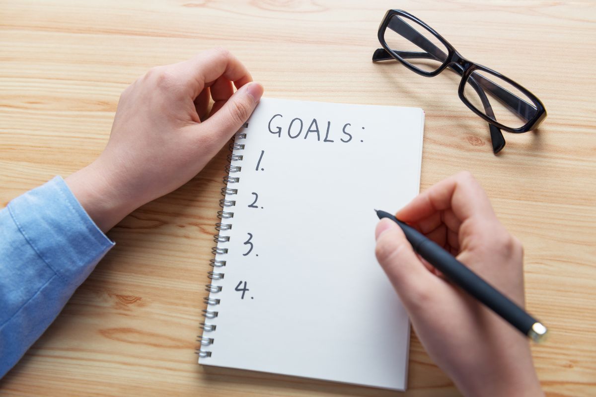 The image of an eyeglass and a girl's hand and writing on the notebook referring to setting a clear goal makes it easier to stay on track and reminds you why saving is worth it.