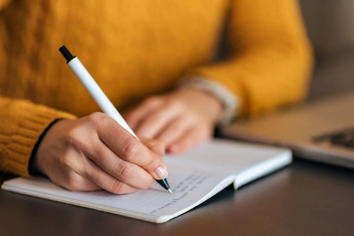 The photo shows a laptop and a girl's hand writing on the notebook which refers to writing and down your affirmations that can change the way you think about money.