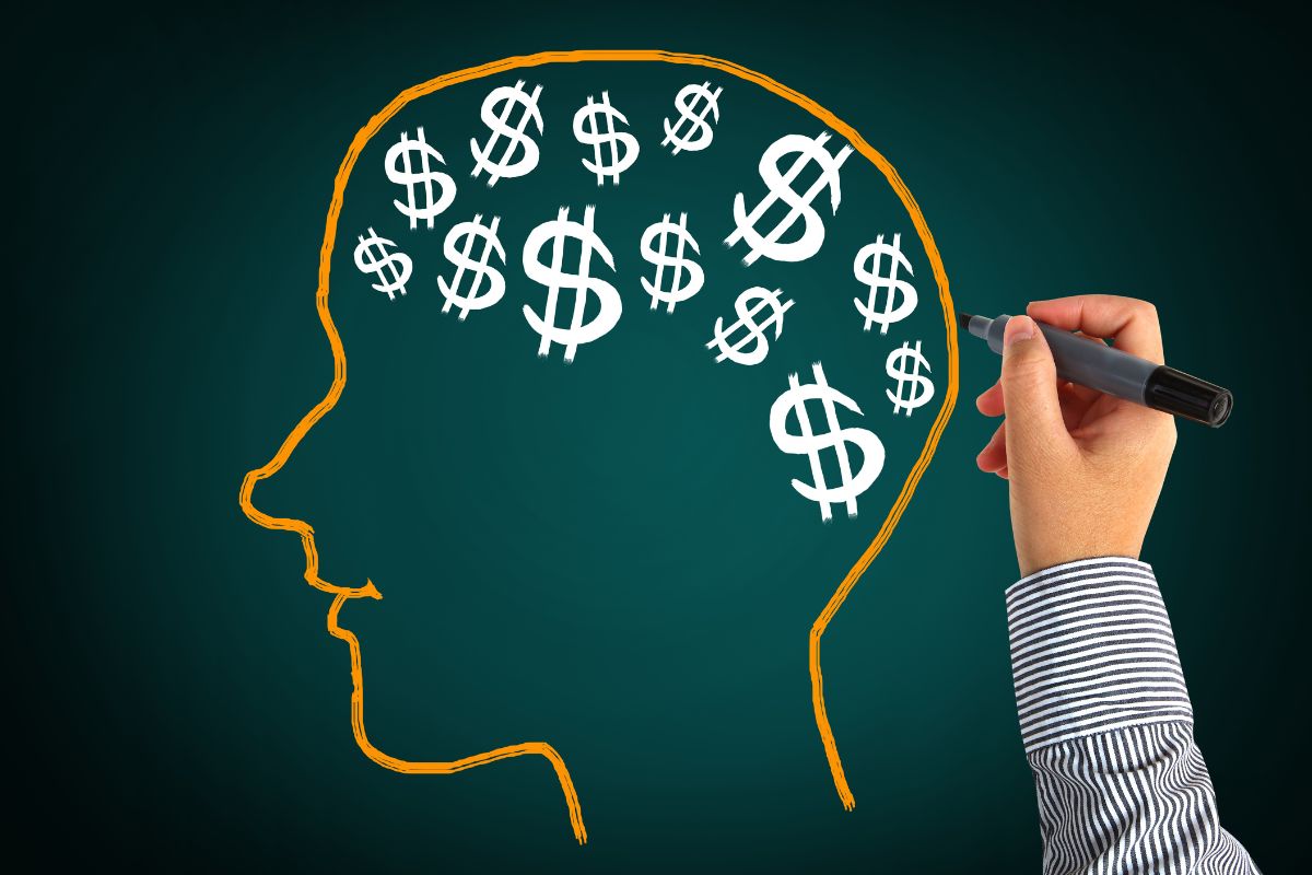 The picture shows a man's hand holding a marker and a board with human drawings and dollar signs referring to letting go of limiting beliefs around money that affects how you manage it.