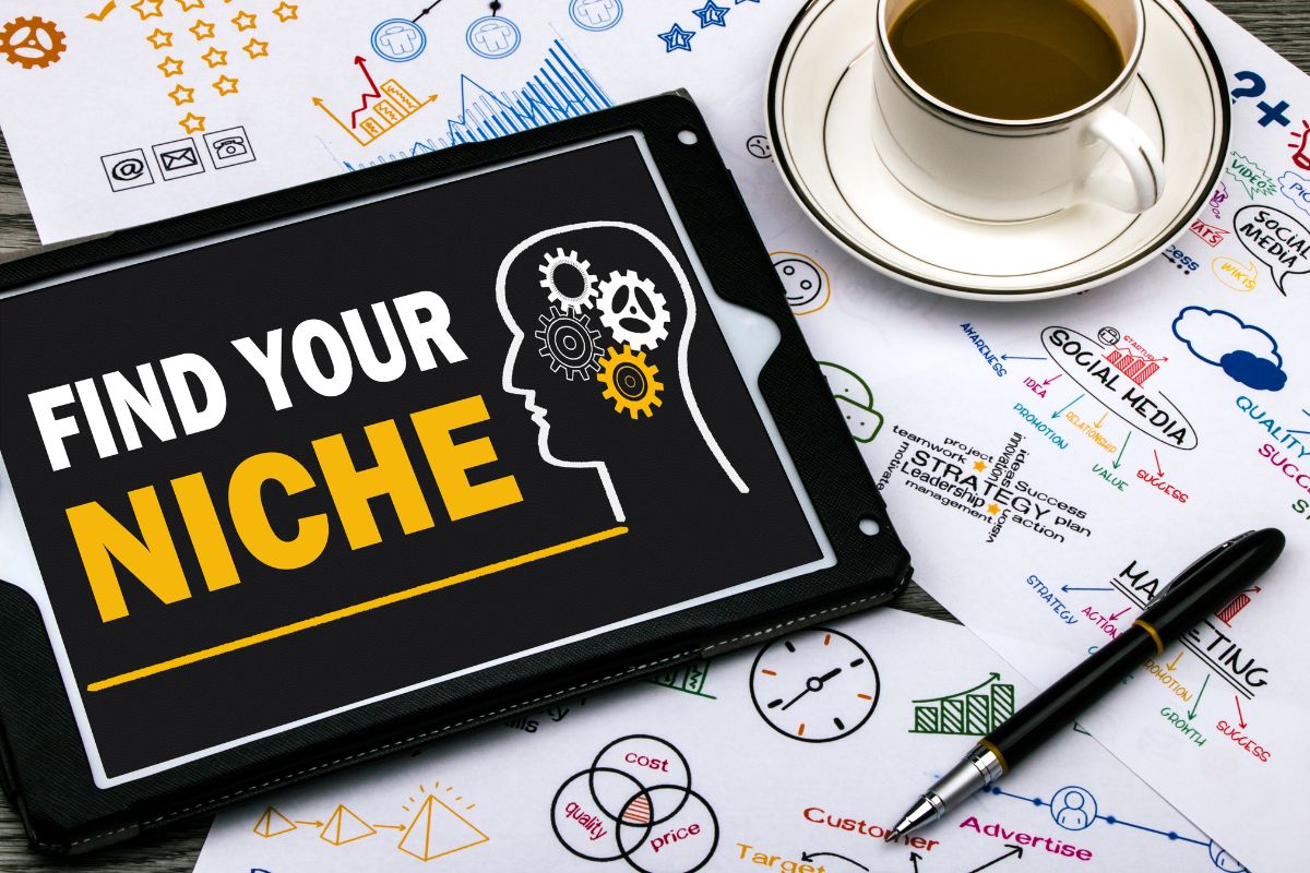 The image tells about papers with notes and graphs, a pen, a coffee, and a tablet device with a screen display referring to choosing a niche that helps you stand out and sell more.