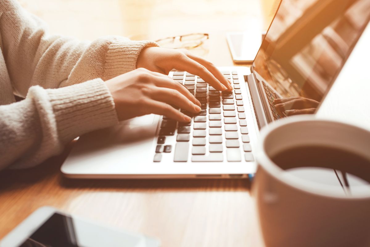 The image shows a coffee, a phone, and a girl's hand using a laptop indicating that selecting a print-on-demand platform can make running your business easier and help you make more sales.