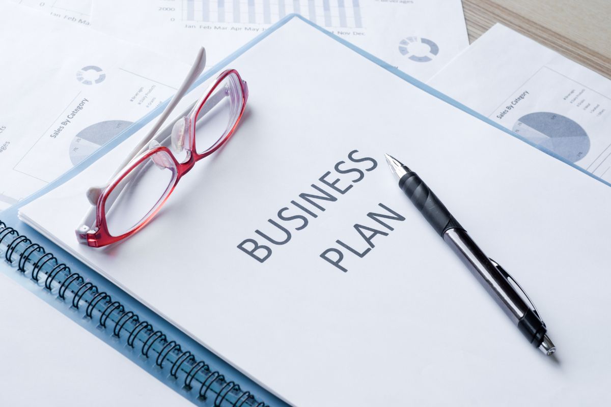 The photo of papers with graphs and notes, a pen, an eyeglass, and a notebook with notes means creating a business plan helps you avoid mistakes and keeps you on track to turn your side hustle into a full-time income.