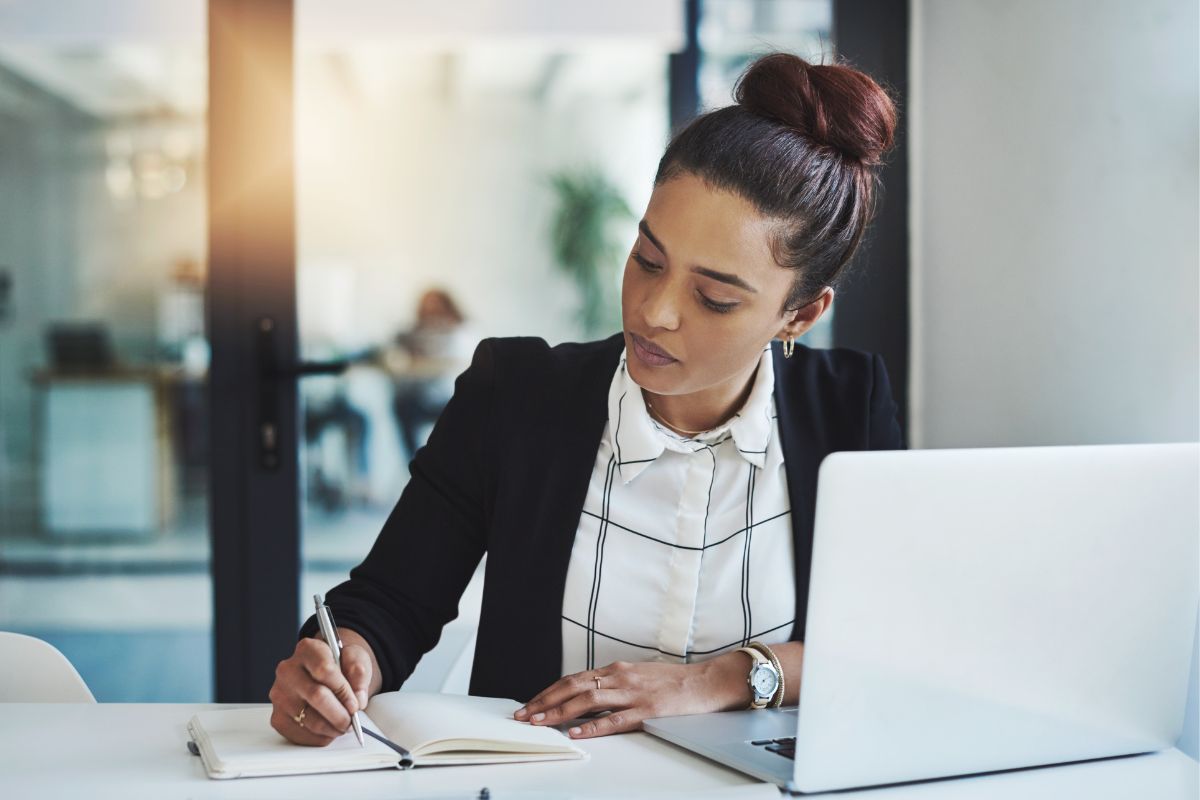The photo shows a laptop and a girl writing in the notebook to stay consistent and adapt to market changes to turn their side hustle into a full-time income.