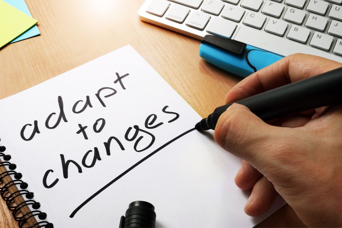 The photo of a keyboard, papers, and a man's hand and writing on the notebook which refers to adaptability and resilience are more likely to find new ways to make money and stay successful.