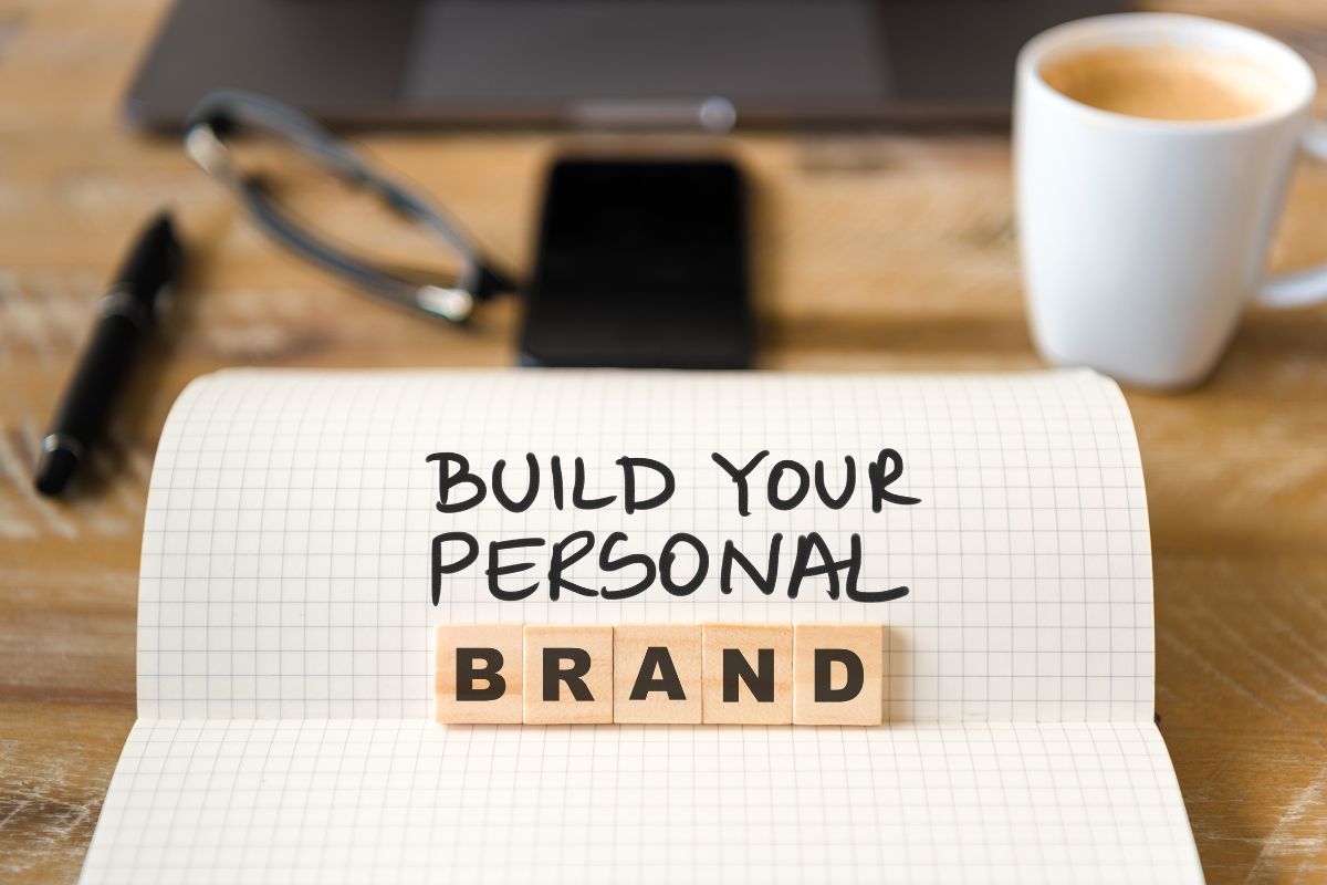 The picture of a laptop, a phone, an eyeglass, a coffee, and a notebook with notes and wooden cubes with letters indicating personal branding helps you stand out and attract better opportunities.