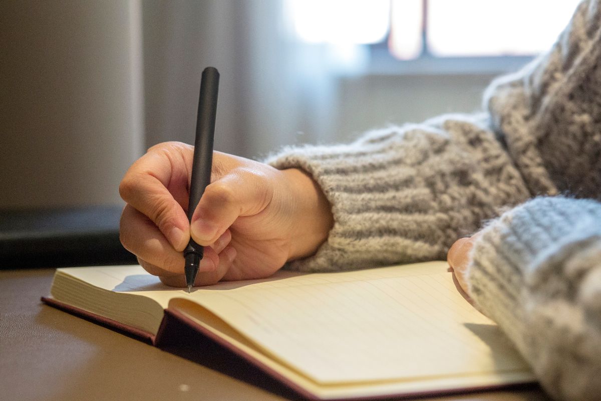 The picture shows a girl's hand and writing on the notebook referring to creating things to be grateful for list can boost your mood and make a no-spend challenge feel less restrictive. 