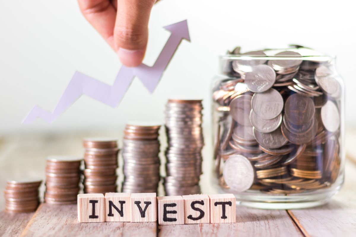 The photo tells about coin stocks, a jar filled with coins, wooden cubes with letters, and a man's hand holding an arrow which means a lifestyle of savings and investing so financial independence becomes possible much faster than most people expect.