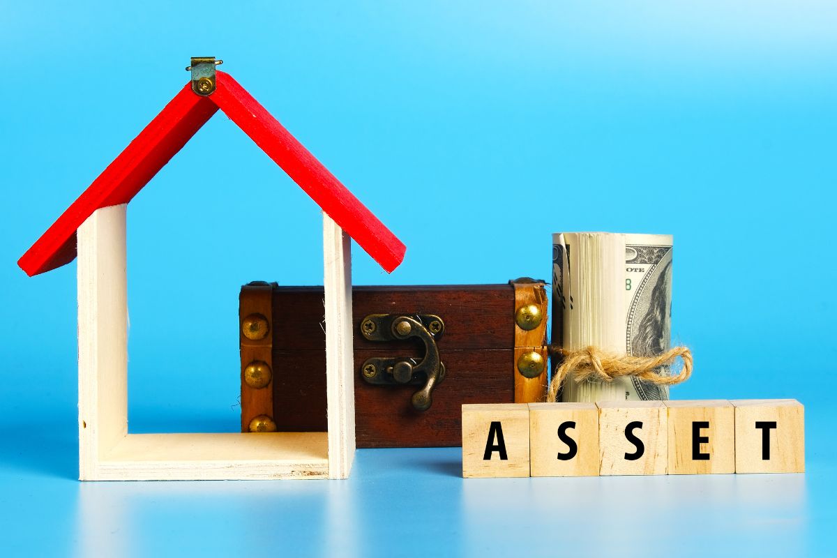 The picture shows a wooden house figure, a wooden box, rolled dollar bills, and wooden cubes with letters which means investing in assets allows savings to grow consistently over time.