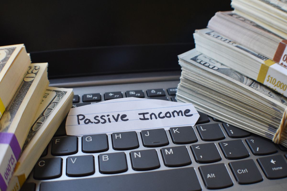 The photo shows a laptop, bucks of dollar bills, and a piece of paper with a note that refers to building passive income that provides money without needing to work.