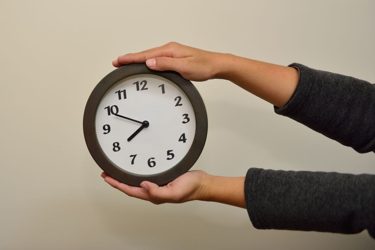 The image shows a girl's hand holding a clock referring to setting a waiting period before non-essential purchases keep money in the bank instead of going to things that don't really matter.