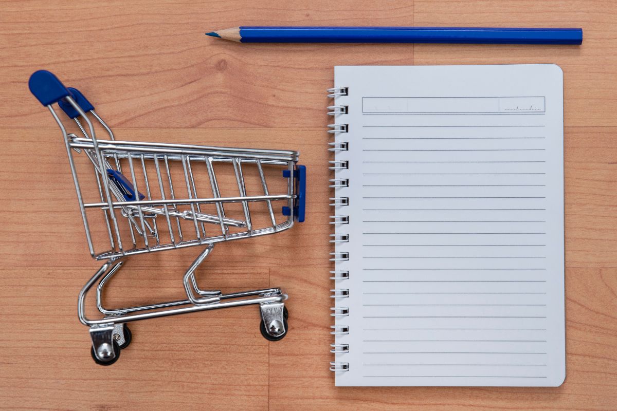The photo shows a pen, a notebook, and a mini cart which refers to creating a specific shopping list before going out preventing impulse purchases, and keeping spending in check.
