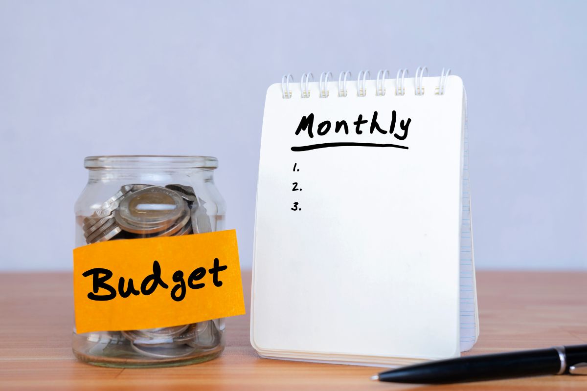 The photo tells about a pen, a notebook with notes, a jar filled with coins, and a note indicating that establishing a monthly fun money budget helps build better spending habits while still enjoying small treats.