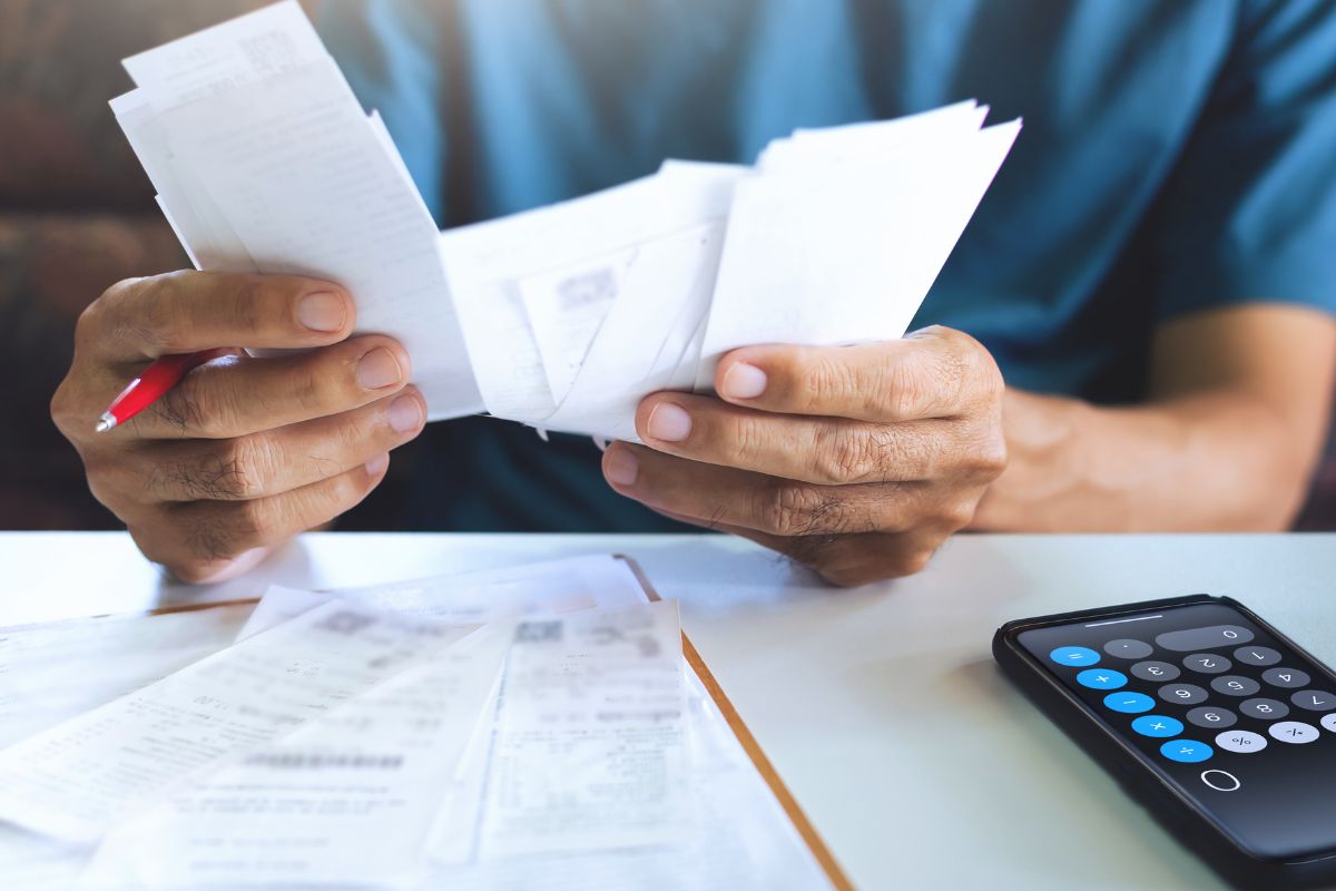 The picture shows a phone with a calculator, receipts on the table, and a man's hand holding and checking receipts referring to reflect on past impulse purchases making it easier to make better choices moving forward.