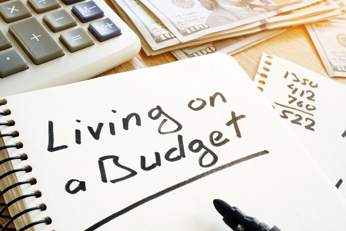 The picture shows dollar bills, a calculator, a marker, and a notebook with notes referring to learning how to live on a budget can take control of your spending and work toward financial stability.