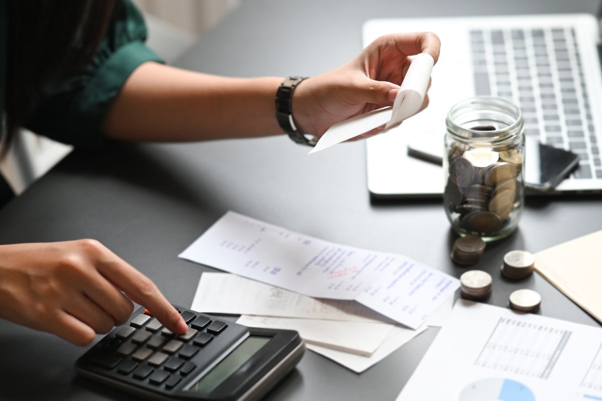 The picture shows a laptop, a jar filled with coins, receipts on the table, papers, and a girl's hand holding a receipt while calculating indicating that tracking your expenses for a month helps you see patterns and know where your money goes.
