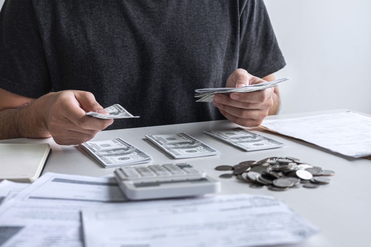 The image tells about papers on the table, a calculator, coins, and a man dividing dollar bills into groups referring to categorize your spending making it clear where you can cut back.