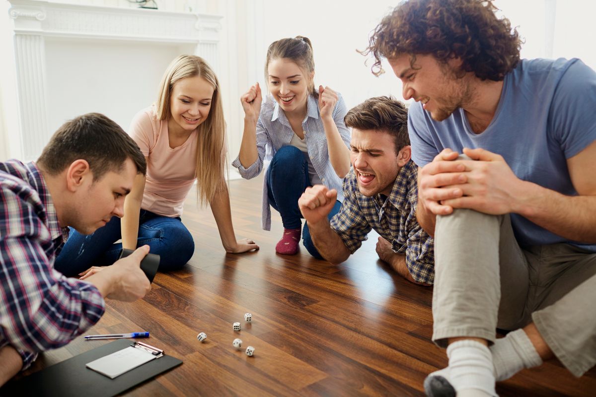The image shows a group of friends playing games with a pen and a clipboard referring to finding alternative ways to cope with stress helps you stop overspending without feeling deprived.
