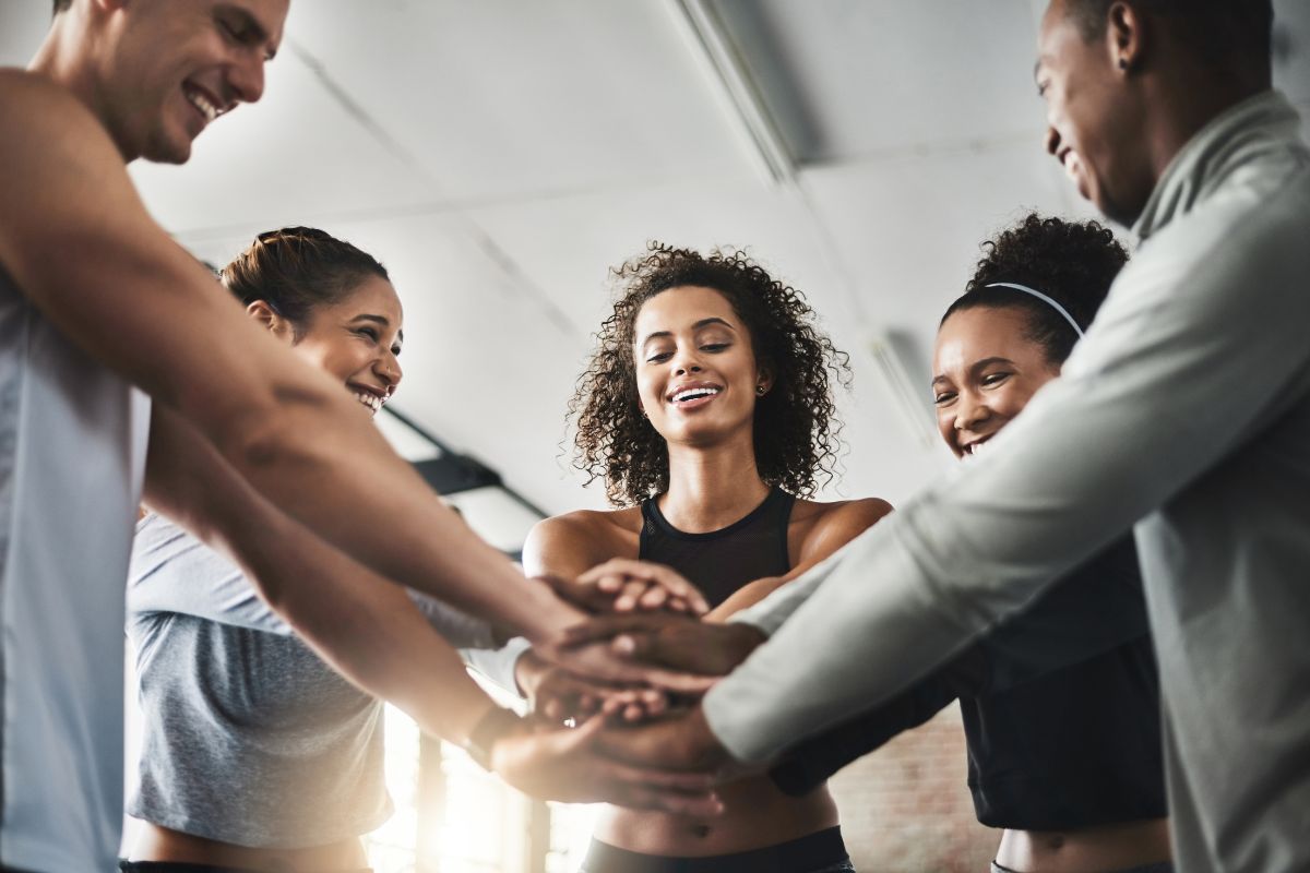 The image shows a group of people joining hands together referring to surrounding yourself with supportive people making it easier to stick to your budget and avoid unnecessary spending.