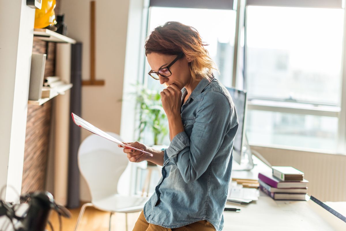 The picture shows books, a chair, a computer set, and a girl looking at the papers which refers to regularly reviewing progress making it easier to stay on track and reach savings goals.